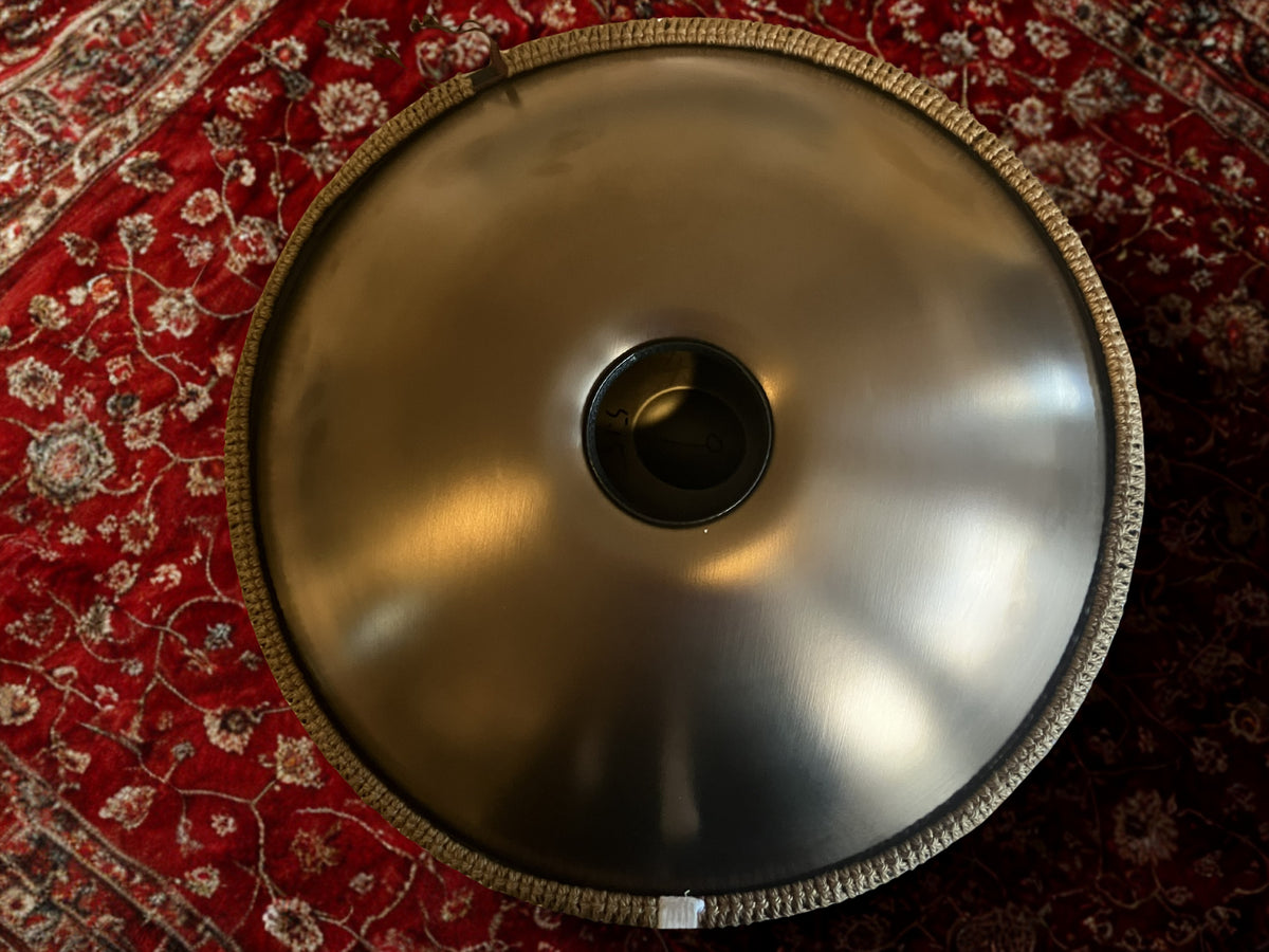 Underside of a gold-toned handpan showing the sound hole on a red patterned rug