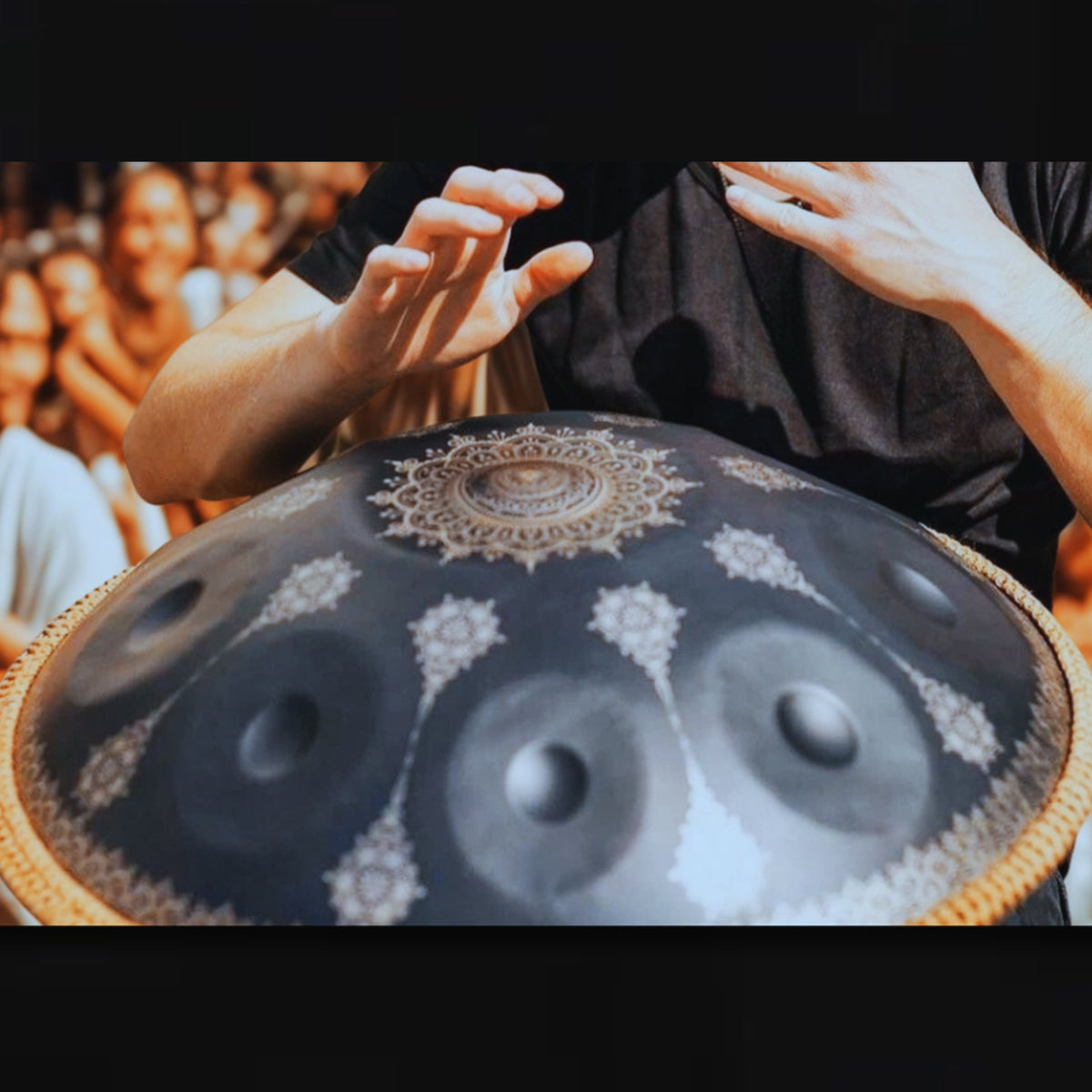 A close-up of a person playing a decorative steel pan drum, with hands raised and the ornate dark metal drum surface in focus against a blurred crowd background. black_yantra_fossey_handpan_closeup_D-kurd_stainless_steel_handpansounds