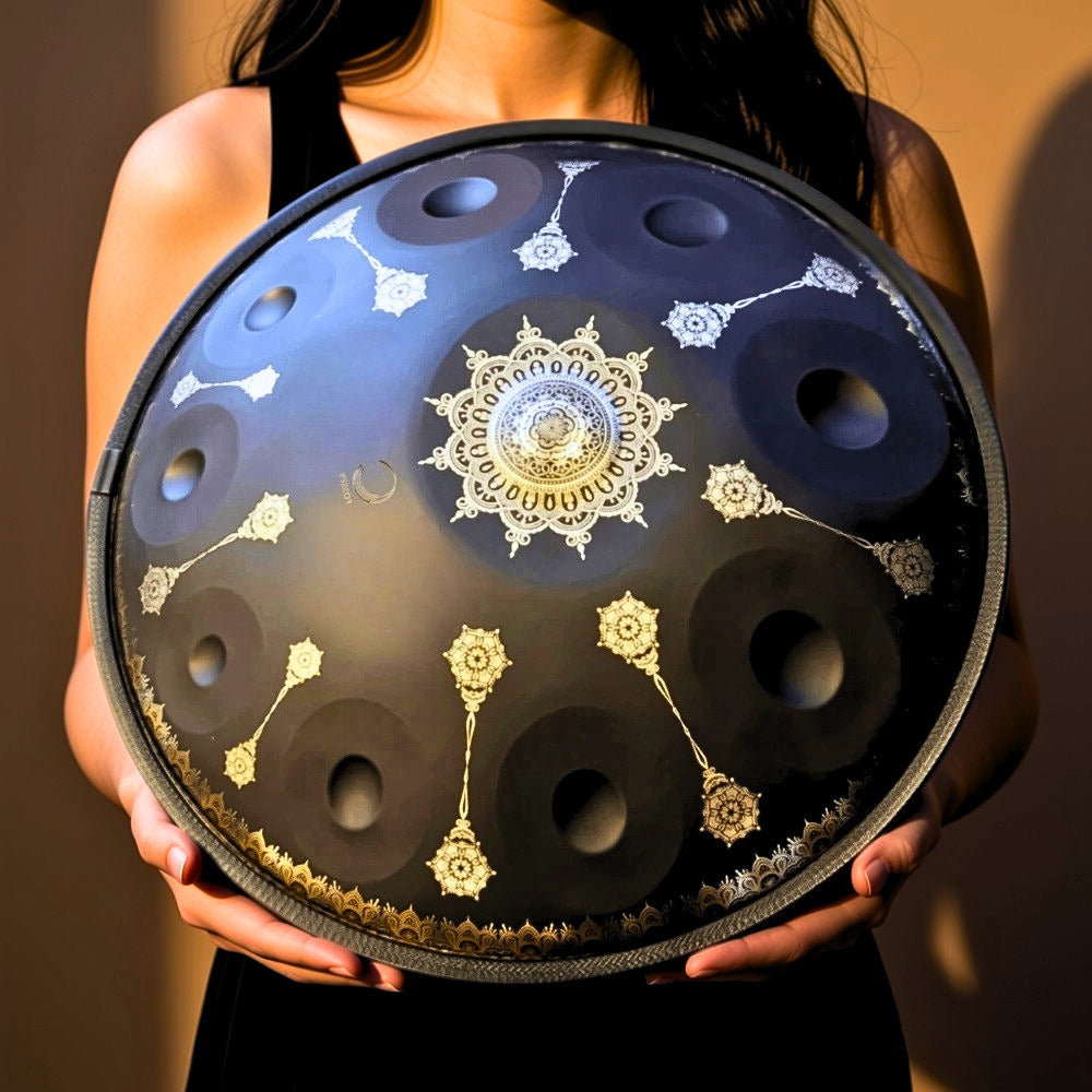 A person holds a large handpan drum, also known as a hang drum, featuring a black metallic body with intricate gold and silver filigree patterns and circular sound holes. Black Yantra Fossey Handpan 10-note, D Kurd, Stainless Steel, 22 inch. handpansounds