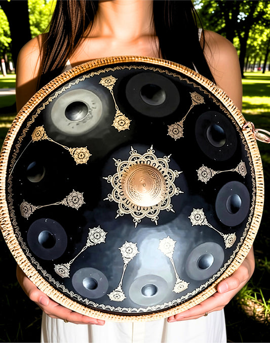A person outdoors holding a large decorative handpan drum with a black interior, gold-toned central medallion and floral motifs, and a light natural wood rim. Black Yantra Fossey Handpan 10-note, D Kurd, Stainless Steel, handpansounds, mandala design