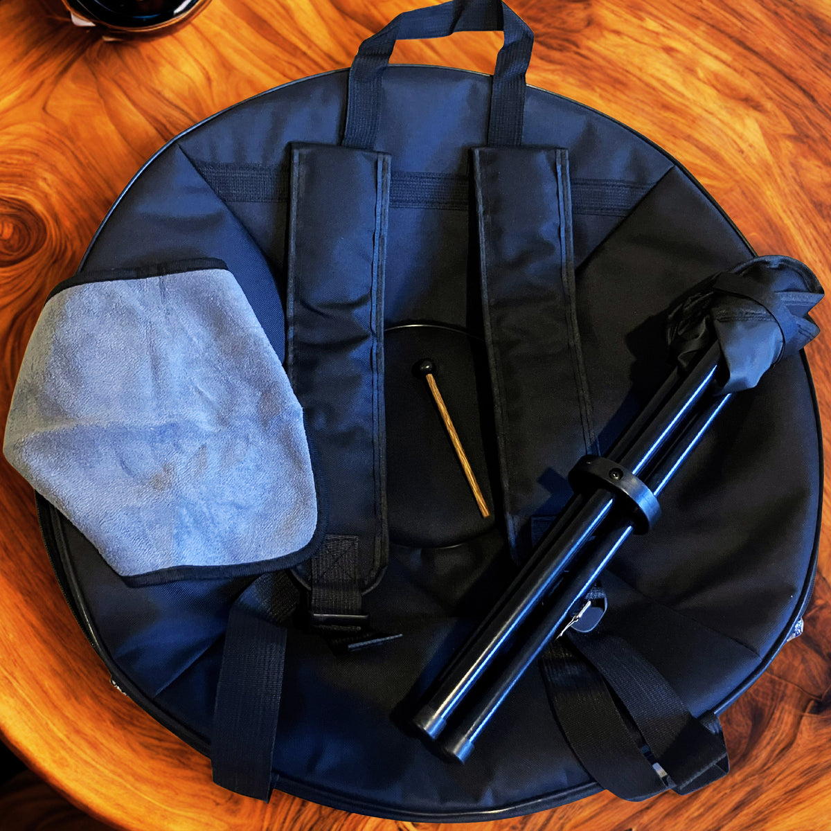 A black circular handpan carrying case laid flat on a wooden table, with a gray cleaning cloth, a mallet, and a folded black handpan standplaced on top. Fossey handpan free accessories with your handpan at handpansounds ... 