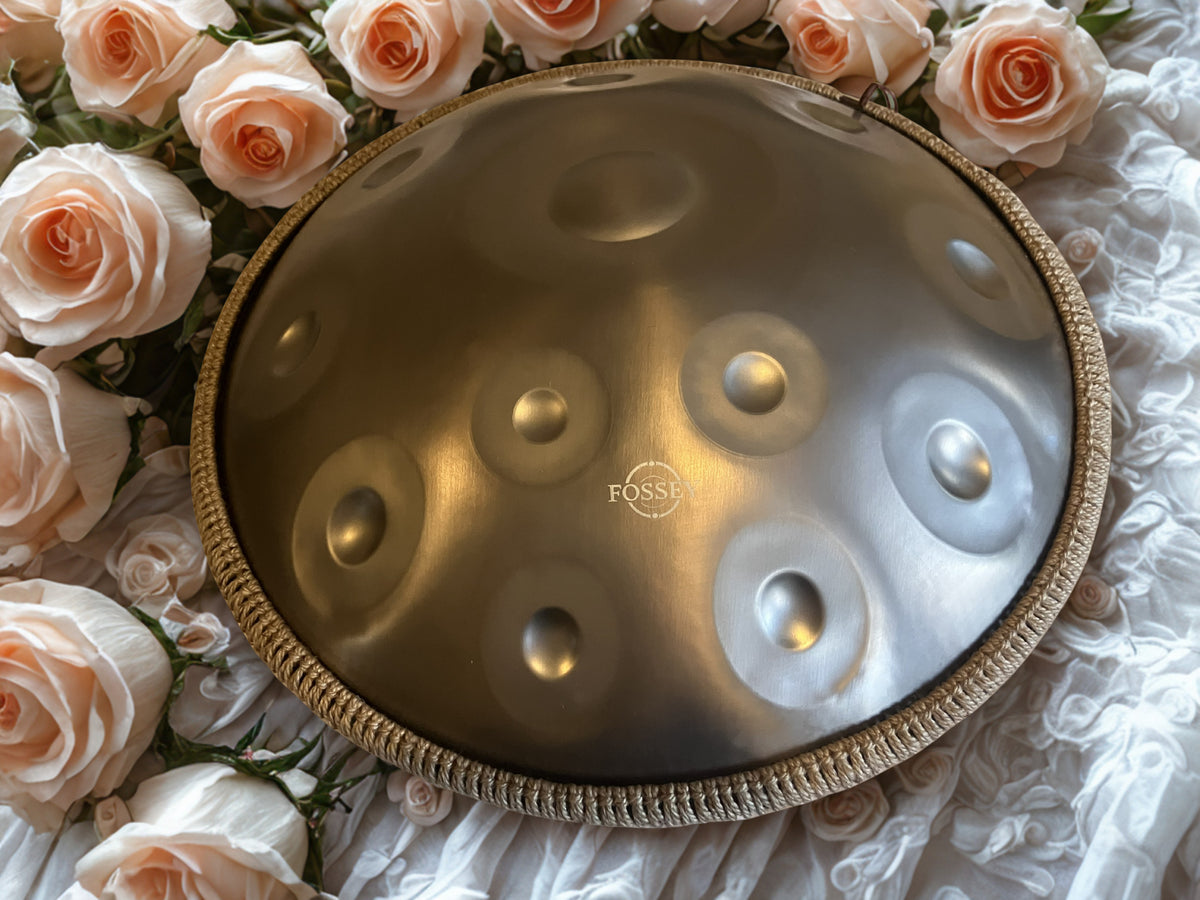 Fossey handpan surrounded by pale roses on white fabric. Gold Standard Fossey Handpan, 12-Note D Kurd, Stainless Steel, 432Hz