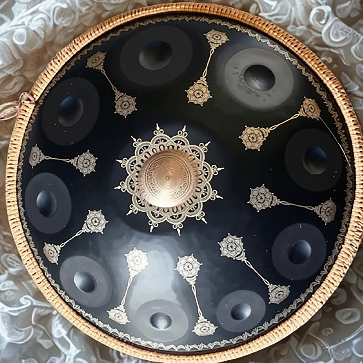 A round decorative tray or platter with a black center, gold-toned filigree motifs, and a woven tan rim, photographed from above on a light patterned fabric background. black_yantra_fossey_handpan_closeup_D-kurd_stainless_steel_handpansounds
