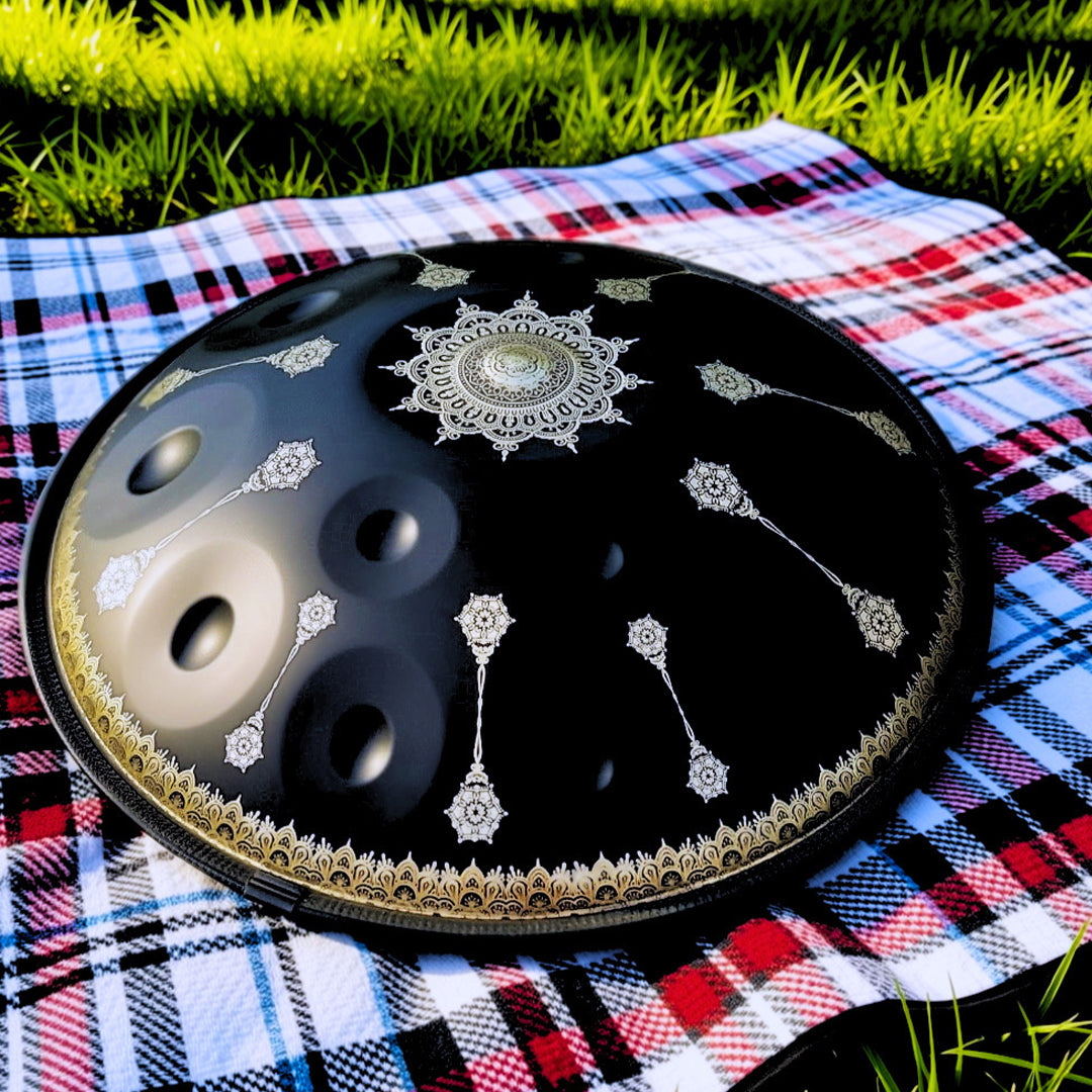 A black handpan drum with ornate silver and gold decorative patterns, resting on a red, white, and blue plaid blanket outdoors on grass.