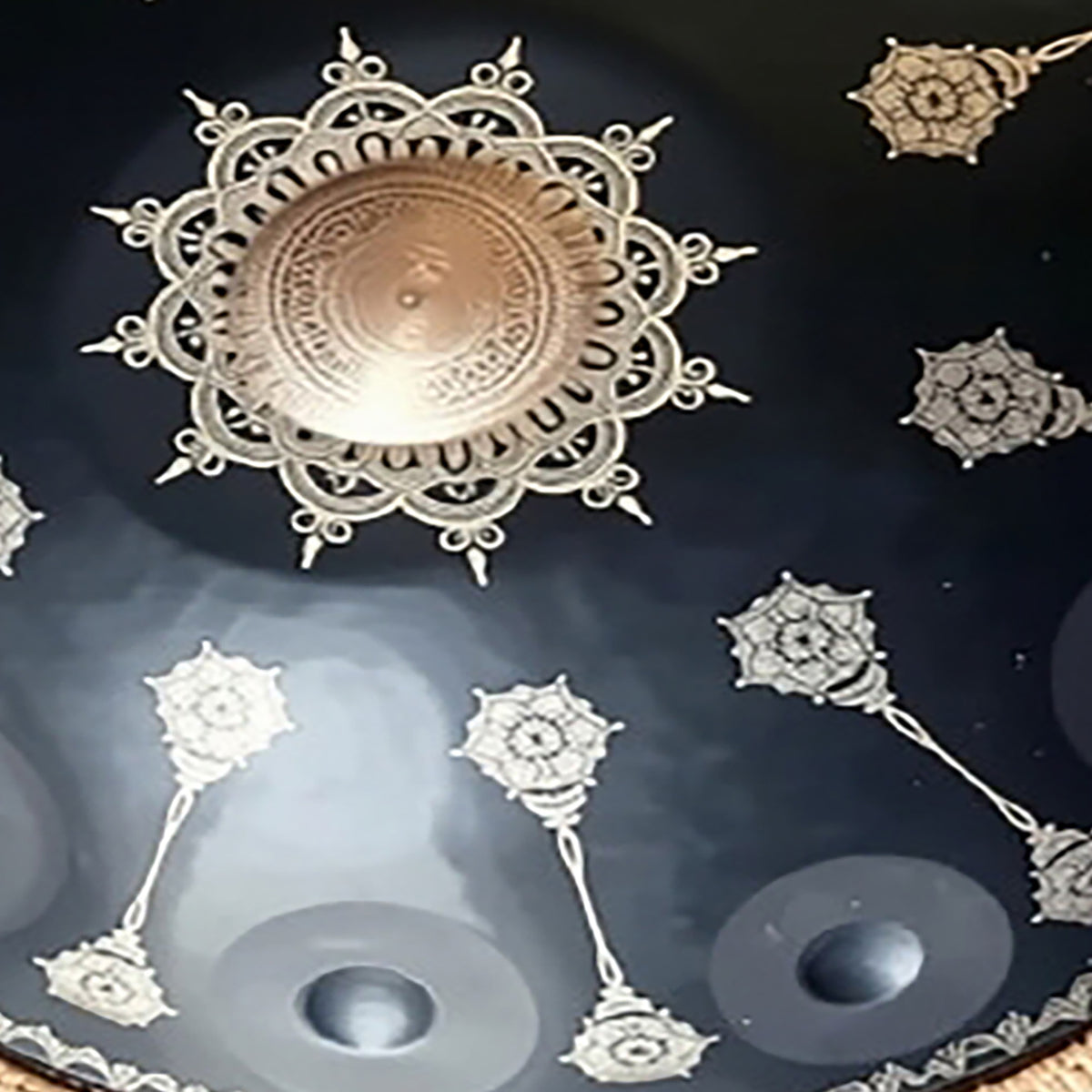 A close-up of a dark round decorative fossey handpan featuring a central metallic gold-toned medallion surrounded by intricate silver filigree patterns and smaller matching motifs.